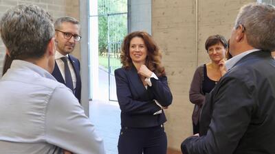 Grup de persones reunides a l’interior de l’edifici de la promoció B2. Al centre, Kata Tütto, vestida amb un vestit blau marí somriu i mira endavant, visiblement involucrada en la conversa. Al costat seu, un home amb ulleres i vestit fosc també sembla estar atent a la interacció. Altres persones estan parcialment visibles a les vores de la imatge.