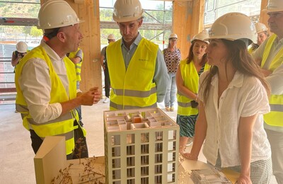 La imatge mostra un grup de persones durant la visita a l’obra. La majoria porten cascos blancs d’obra i armilles reflectants grogues i estan reunides al voltant d’una maqueta d’un edifici d’apartaments de diversos pisos. L’entorn és de construcció.