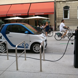 Cotxe elèctric carregant-se en un electrolinera i al fons una jove en bicicleta elèctrica