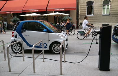 Cotxe elèctric carregant-se en un electrolinera i al fons una jove en bicicleta elèctrica