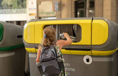Foto: Martí Petit Dona al carrer davant un gran contenidor de reciclatge de color groc depositant una ampolla de plàstic