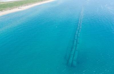 Vista aèria d´una extensa línia d´estructures geotubs submergides en aigües de color turquesa. A la part superior de la imatge es veu una platja de sorra amb vegetació.