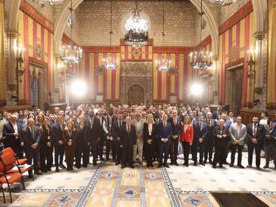 Foto de grup Participants al WMS posant en la foto de grup dins del Saló de Cent. L´interior, d’origen medieval, compta amb alts sostres de pedra, canelobres elaborats i cortines de color vermell i groc. El pis també està decorat amb un disseny de rajoles intricat. Les persones del grup vesteixen una varietat de vestits formals.