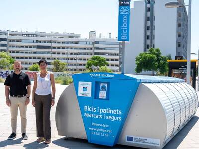 Un home i una dona, dempeus davant d'un Bicibox durant la presentació de la nova estació. El Bicibox és un contenidor metàl·lic de color blau i platejat, amb un panell digital a la part frontal. El lloc és una amplia plaça pública sense zona verda.