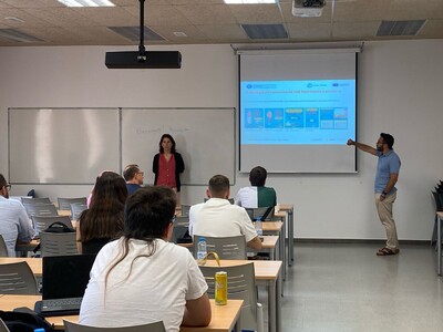Aula de formació on hi ha dos docents, una dona dreta davant una pissarra i un home que senyala una imatge en una pantalla de projecció. Hi ha diverses persones assegudes a les taules de l’aula