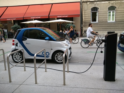 Cotxe elèctric carregant-se en un electrolinera i al fons una jove en bicicleta elèctrica