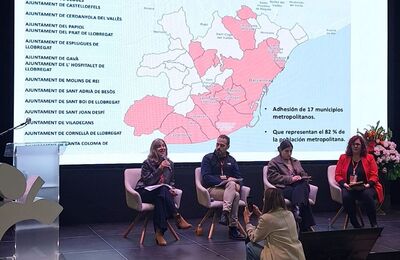 Taula rodona on quatre persones estan assegudes en cadires, participant a la conversa. Darrere seu, es projecta una presentació en una pantalla gran que mostra un mapa de municipis de l’àrea metropolitana de Barcelona. Elisabet Tejero, en primer pla, està usant un micròfon fent la seva ponència. Davant seu, una dona li fa una fotografia.