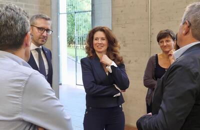 Grup de persones reunides a l’interior de l’edifici de la promoció B2. Al centre, Kata Tütto, vestida amb un vestit blau marí somriu i mira endavant, visiblement involucrada en la conversa. Al costat seu, un home amb ulleres i vestit fosc també sembla estar atent a la interacció. Altres persones estan parcialment visibles a les vores de la imatge.
