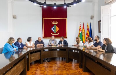 La imatge mostra la reunió formal al voltant d’una taula en forma de U. Hi ha deu persones assegudes, cinc a cada costat, en la sala de juntes de l’ajuntament amb un estendard darrere de la taula, i banderes al fons a la dreta de la imatge. L’ambient és seriós i formal.