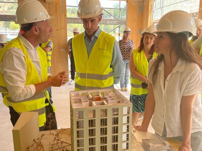 La imatge mostra un grup de persones durant la visita a l’obra. La majoria porten cascos blancs d’obra i armilles reflectants grogues i estan reunides al voltant d’una maqueta d’un edifici d’apartaments de diversos pisos. L’entorn és de construcció.